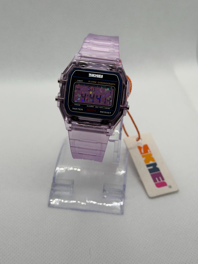 Puede incluir: Un reloj digital lavanda transparente con una esfera negra que muestra la hora. El reloj tiene una correa lavanda a juego y est&aacute; sobre un soporte de pl&aacute;stico transparente. La marca "SKMEI" es visible en la esfera y la etiqueta.