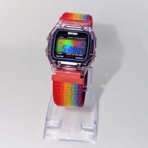 May include: A digital wristwatch with a transparent purple plastic case and a rainbow-striped fabric strap. The watch face displays the brand name SKMEI, the text ALARM CHRONOGRAPH, and WATER 50M RESIST. The digital screen shows the time 5:08:04.