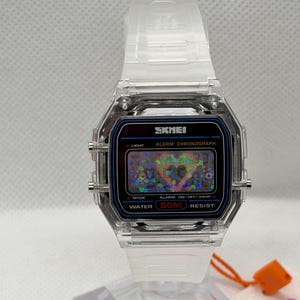 May include: A digital wristwatch with a clear, transparent plastic case and strap. The rectangular watch face features the brand name SKMEI, the text ALARM CHRONOGRAPH, and WATER 50M RESIST. The digital display shows a holographic heart graphic.