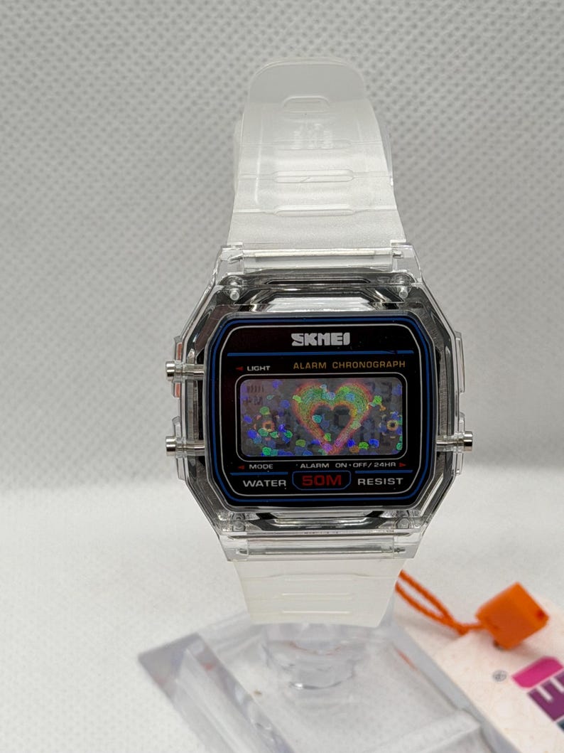 May include: A digital wristwatch with a clear, transparent plastic case and strap. The rectangular watch face features the brand name SKMEI, the text ALARM CHRONOGRAPH, and WATER 50M RESIST. The digital display shows a holographic heart graphic.