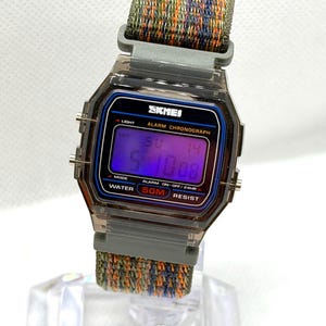 May include: A digital wristwatch with a translucent dark gray case and a rectangular face. The display shows the text SKMEI, ALARM CHRONOGRAPH, and WATER 50M RESIST. It features a woven fabric strap with a multicolored pattern of green, orange, and blue.