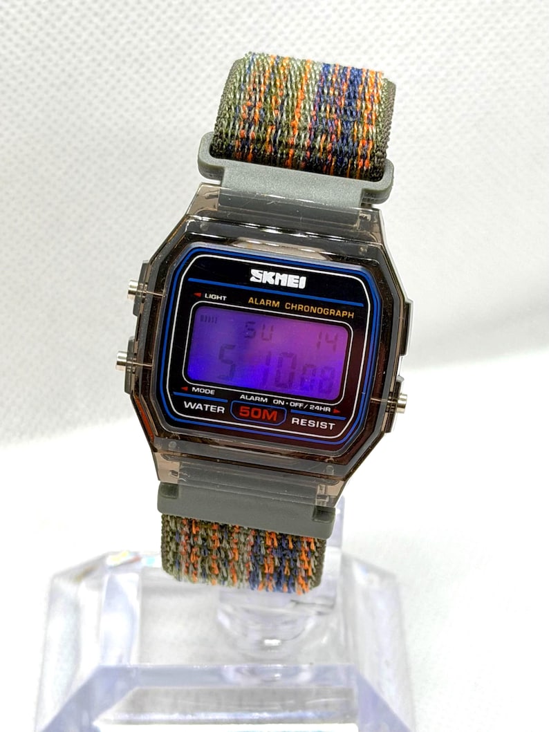 May include: A digital wristwatch with a translucent dark gray case and a rectangular face. The display shows the text SKMEI, ALARM CHRONOGRAPH, and WATER 50M RESIST. It features a woven fabric strap with a multicolored pattern of green, orange, and blue.