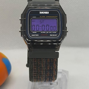 May include: A digital wristwatch with a dark gray rectangular case and a purple digital display. The watch face features the text SKMEI, ALARM CHRONOGRAPH, and WATER 50M RESIST. It has a textured, multi-colored fabric strap in shades of black, blue, and orange.