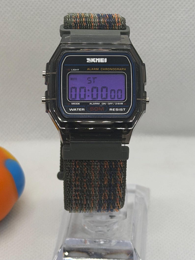 May include: A digital wristwatch with a dark gray rectangular case and a purple digital display. The watch face features the text SKMEI, ALARM CHRONOGRAPH, and WATER 50M RESIST. It has a textured, multi-colored fabric strap in shades of black, blue, and orange.