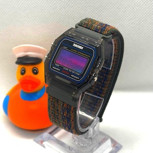 May include: A digital wristwatch with a translucent black plastic case and a multi-colored woven fabric strap. The watch face features the brand name SKMEI, the text ALARM CHRONOGRAPH, and WATER 50M RESIST. The display has a purple gradient tint.