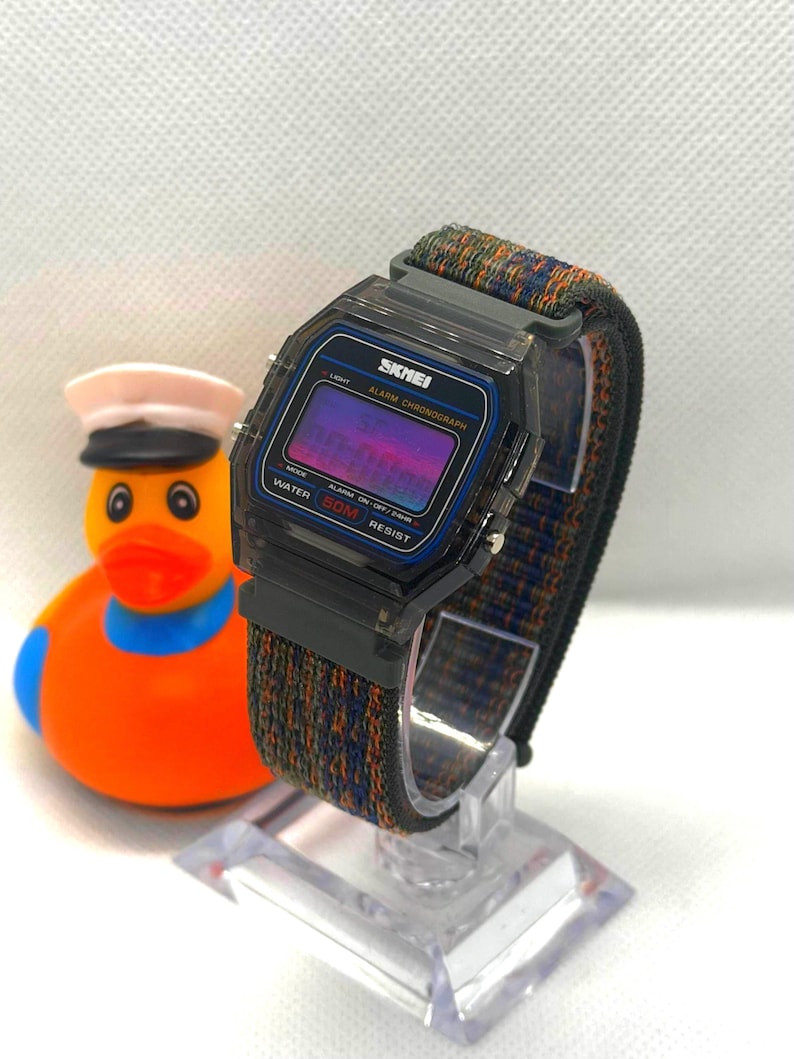 May include: A digital wristwatch with a translucent black plastic case and a multi-colored woven fabric strap. The watch face features the brand name SKMEI, the text ALARM CHRONOGRAPH, and WATER 50M RESIST. The display has a purple gradient tint.