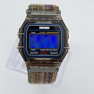 May include: A digital wristwatch with a translucent dark gray plastic case and a blue digital display. The watch face features the text SKMEI, ALARM CHRONOGRAPH, and WATER 50M RESIST. It has a textured, multi-colored fabric strap in shades of green, blue, and orange.