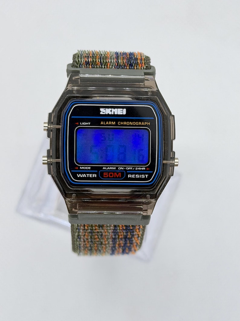 May include: A digital wristwatch with a translucent dark gray plastic case and a blue digital display. The watch face features the text SKMEI, ALARM CHRONOGRAPH, and WATER 50M RESIST. It has a textured, multi-colored fabric strap in shades of green, blue, and orange.
