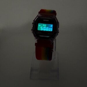 May include: Digital watch with a rainbow-colored strap. The watch face displays the time in a bright green font. The watch has a clear, transparent case and is displayed on a clear stand.