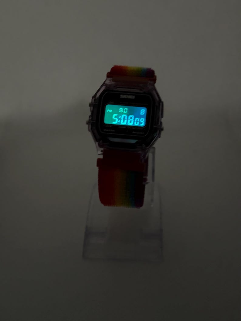 May include: Digital watch with a rainbow-colored strap. The watch face displays the time in a bright green font. The watch has a clear, transparent case and is displayed on a clear stand.