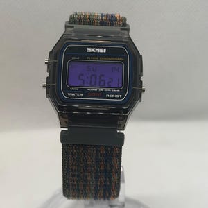 May include: A digital wristwatch with a translucent dark gray octagonal case and a textured fabric strap in shades of blue, green, and orange. The watch face displays the brand name SKMEI, ALARM CHRONOGRAPH, and WATER 50M RESIST text.