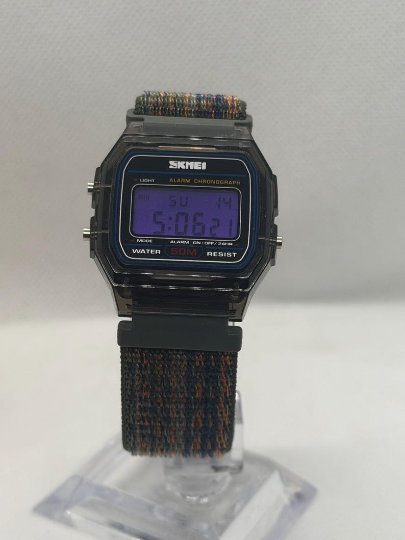 May include: A digital wristwatch with a translucent dark gray octagonal case and a textured fabric strap in shades of blue, green, and orange. The watch face displays the brand name SKMEI, ALARM CHRONOGRAPH, and WATER 50M RESIST text.