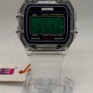 K&ouml;nnte beinhalten: Transparente Digitaluhr mit wei&szlig;em Armband. Das Zifferblatt zeigt die Uhrzeit 3:57 an, mit dem Markennamen "SKMEI". Funktionen: Licht, Alarm und Wasserdichtigkeit bis 50 Meter. Die Uhr wird auf einem transparenten St&auml;nder pr&auml;sentiert.
