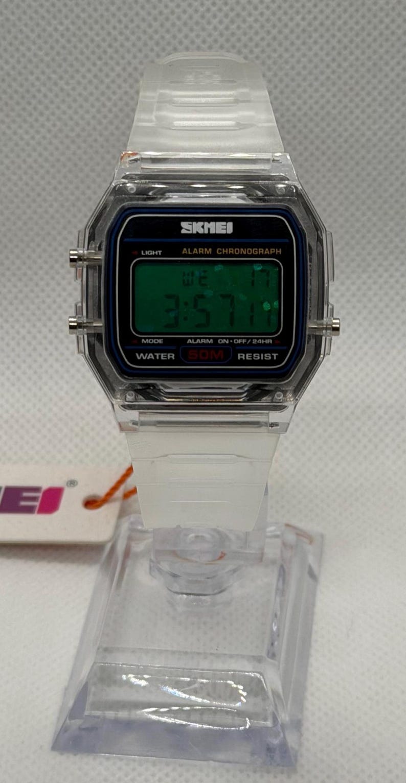 K&ouml;nnte beinhalten: Transparente Digitaluhr mit wei&szlig;em Armband. Das Zifferblatt zeigt die Uhrzeit 3:57 an, mit dem Markennamen "SKMEI". Funktionen: Licht, Alarm und Wasserdichtigkeit bis 50 Meter. Die Uhr wird auf einem transparenten St&auml;nder pr&auml;sentiert.