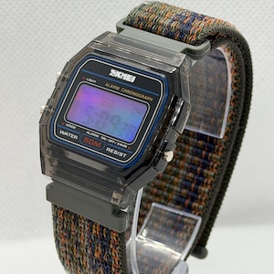 May include: A digital wristwatch with a translucent dark gray plastic case and a rectangular face. The display shows the text SKMEI, ALARM CHRONOGRAPH, and WATER 50M RESIST. It features a woven fabric strap with a multicolored pattern of orange, blue, and green.