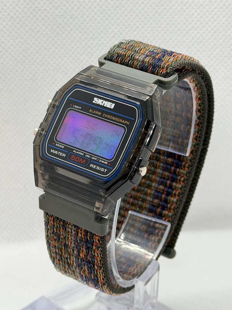 May include: A digital wristwatch with a translucent dark gray plastic case and a rectangular face. The display shows the text SKMEI, ALARM CHRONOGRAPH, and WATER 50M RESIST. It features a woven fabric strap with a multicolored pattern of orange, blue, and green.