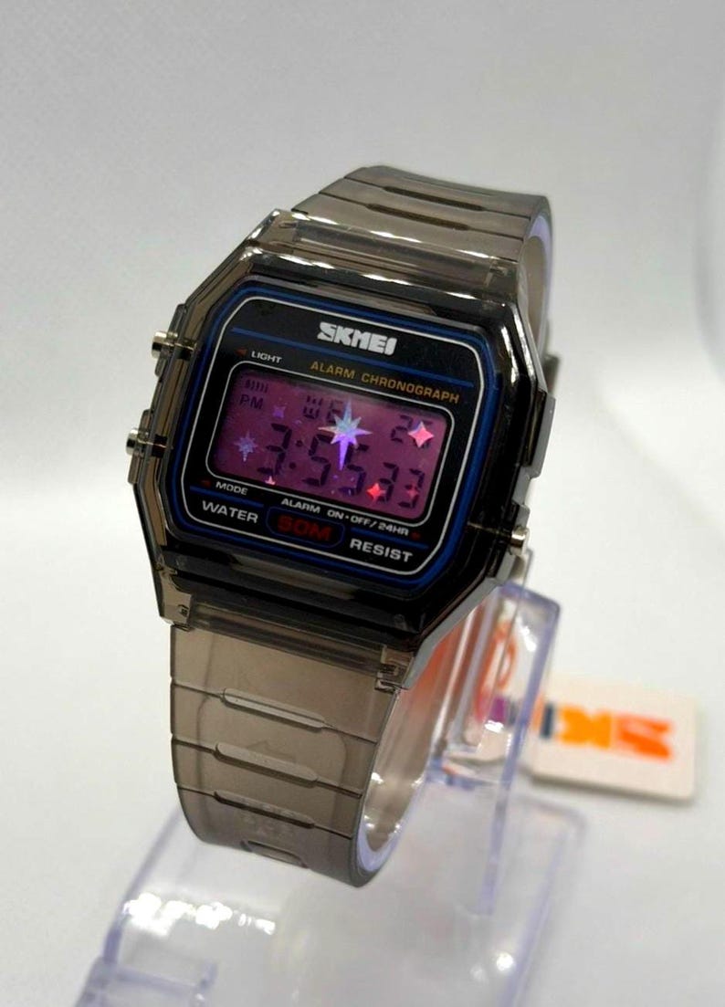 May include: A digital wristwatch with a translucent dark grey resin case and matching link strap. The rectangular face features a black border, the text SKMEI, ALARM CHRONOGRAPH, and WATER 50M RESIST, with a purple digital display showing star icons.