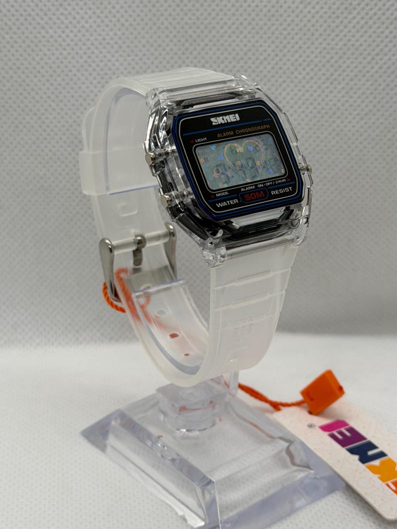 May include: A digital wristwatch with a clear, transparent resin case and matching translucent white silicone strap. The rectangular watch face features a black border, a digital display with the text SKMEI, ALARM CHRONOGRAPH, and WATER 50M RESIST.