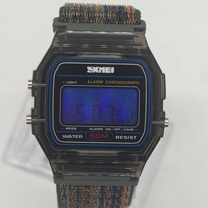 May include: A digital wristwatch with a translucent black plastic case and a blue-tinted display screen. The watch face features the text SKMEI, ALARM CHRONOGRAPH, and WATER 50M RESIST. It includes a textured, multi-colored fabric strap.