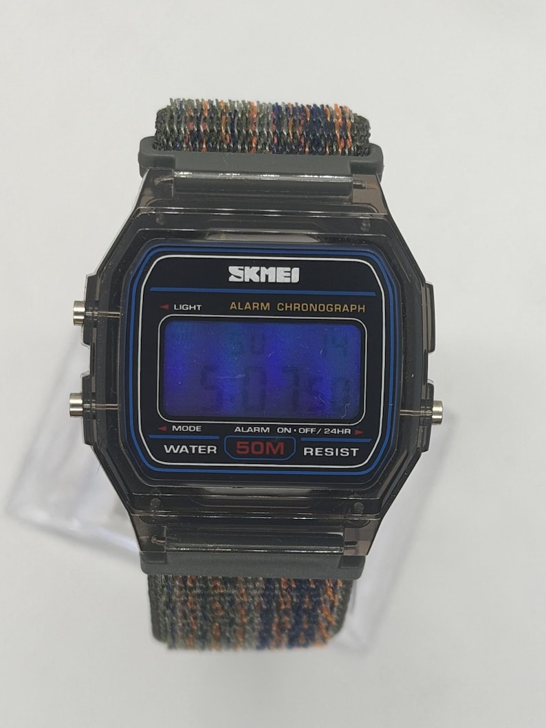 May include: A digital wristwatch with a translucent black plastic case and a blue-tinted display screen. The watch face features the text SKMEI, ALARM CHRONOGRAPH, and WATER 50M RESIST. It includes a textured, multi-colored fabric strap.