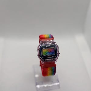 May include: A digital watch with a clear, rectangular case and a rainbow-colored band. The watch face displays the time and other functions. The band is red with a rainbow stripe. The watch is displayed on a clear stand.