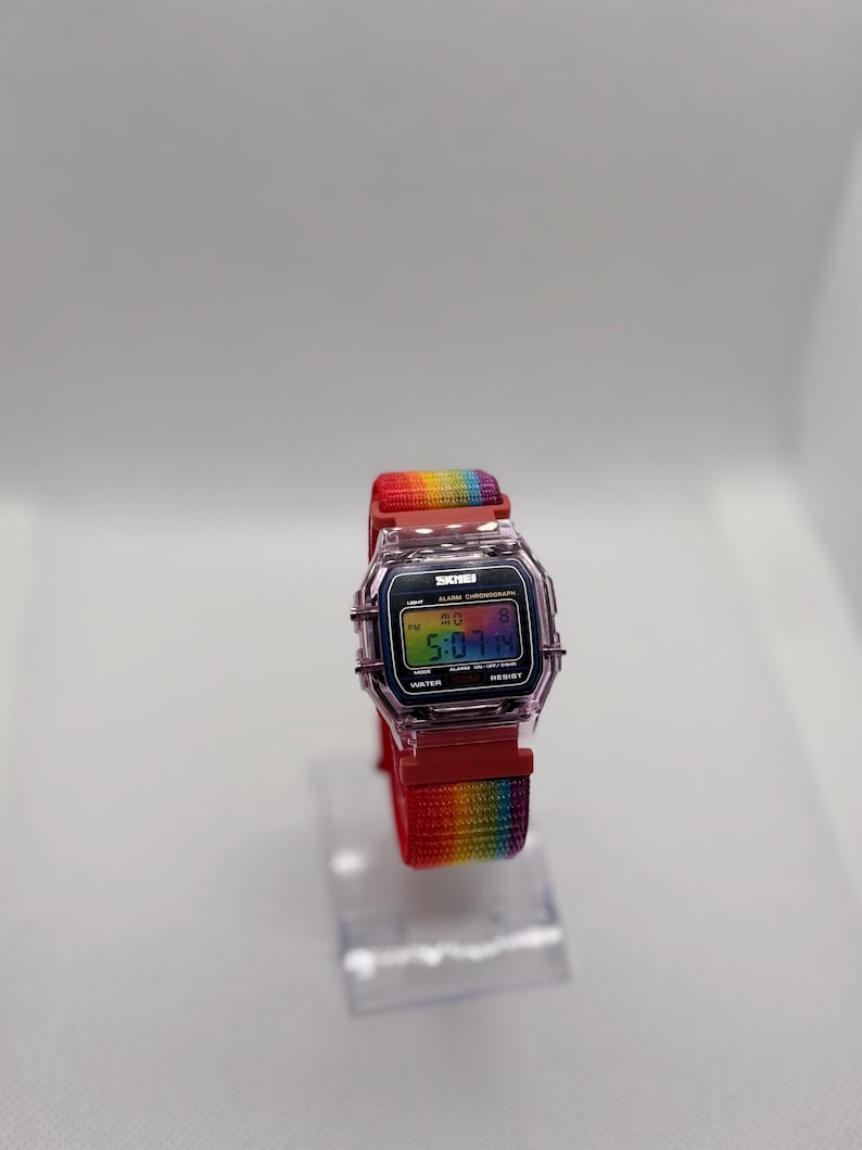 May include: A digital watch with a clear, rectangular case and a rainbow-colored band. The watch face displays the time and other functions. The band is red with a rainbow stripe. The watch is displayed on a clear stand.