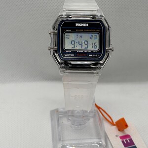 May include: A digital wristwatch with a clear, transparent plastic case and strap. The rectangular watch face features a black border with the text SKMEI, LIGHT, ALARM CHRONOGRAPH, MODE, and WATER 50M RESIST. The digital display shows 9:49 16.