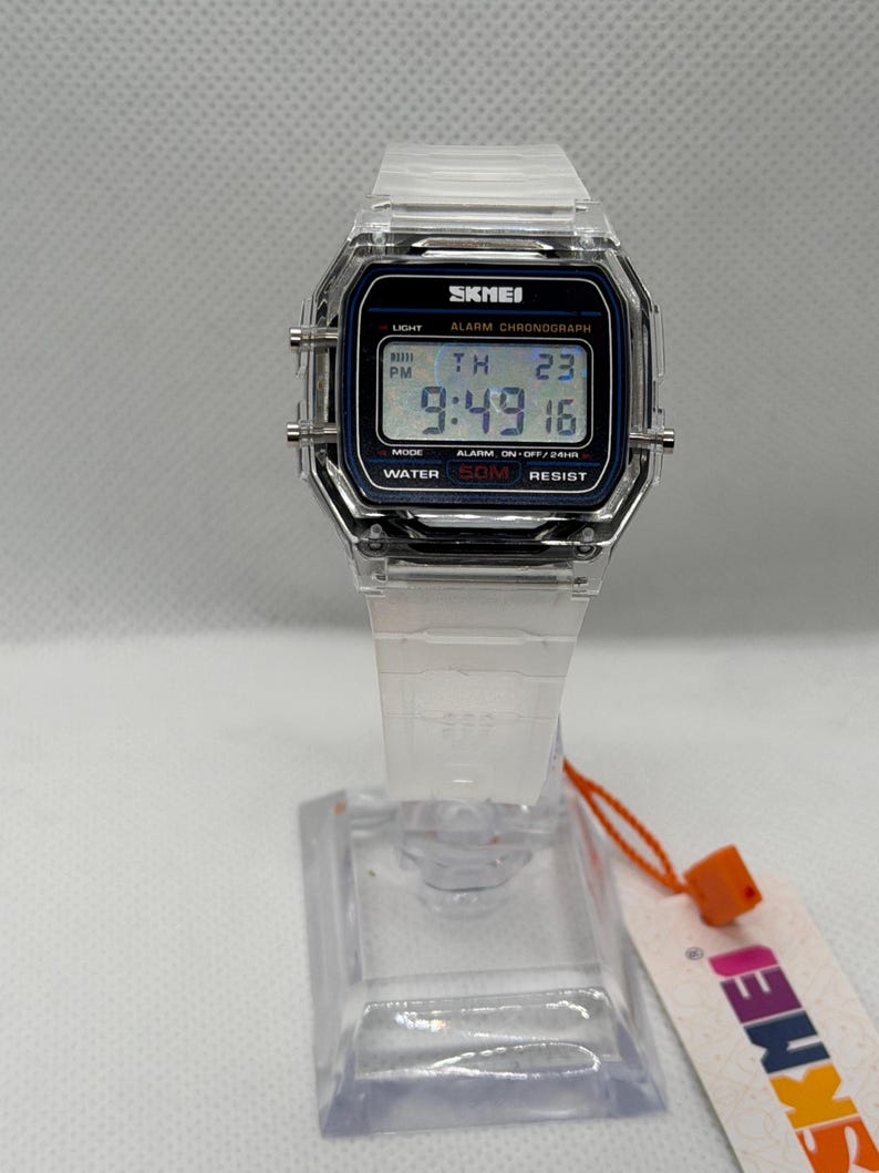 May include: A digital wristwatch with a clear, transparent plastic case and strap. The rectangular watch face features a black border with the text SKMEI, LIGHT, ALARM CHRONOGRAPH, MODE, and WATER 50M RESIST. The digital display shows 9:49 16.