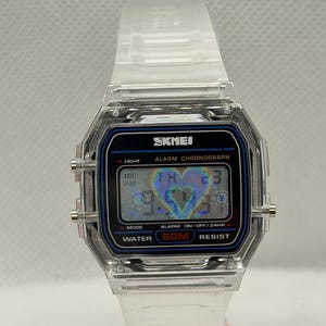 May include: A digital wristwatch with a clear, transparent resin case and strap. The rectangular face features a black border with blue accents, displaying the text SKMEI, ALARM CHRONOGRAPH, and WATER 50M RESIST. A holographic heart icon is visible.