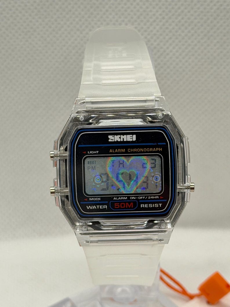 May include: A digital wristwatch with a clear, transparent resin case and strap. The rectangular face features a black border with blue accents, displaying the text SKMEI, ALARM CHRONOGRAPH, and WATER 50M RESIST. A holographic heart icon is visible.