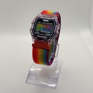 Könnte beinhalten: Eine Digitaluhr mit einem Regenbogenarmband. Das Zifferblatt zeigt die Uhrzeit 5:07:30 an. Die Uhr hat ein transparentes Kunststoffgehäuse und einen roten Verschluss. Das Armband ist ein lebendiger Mix aus Regenbogenfarben.