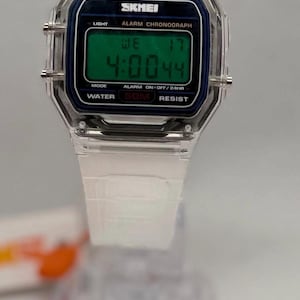 K&ouml;nnte beinhalten: Transparente Digitaluhr mit rechteckigem Zifferblatt. Die Uhr zeigt die Zeit 4:00:44 auf einem gr&uuml;nen Bildschirm an. Sie hat ein transparentes Kunststoffarmband und ein schwarz-blaues Zifferblatt mit der Marke "SKMEI".
