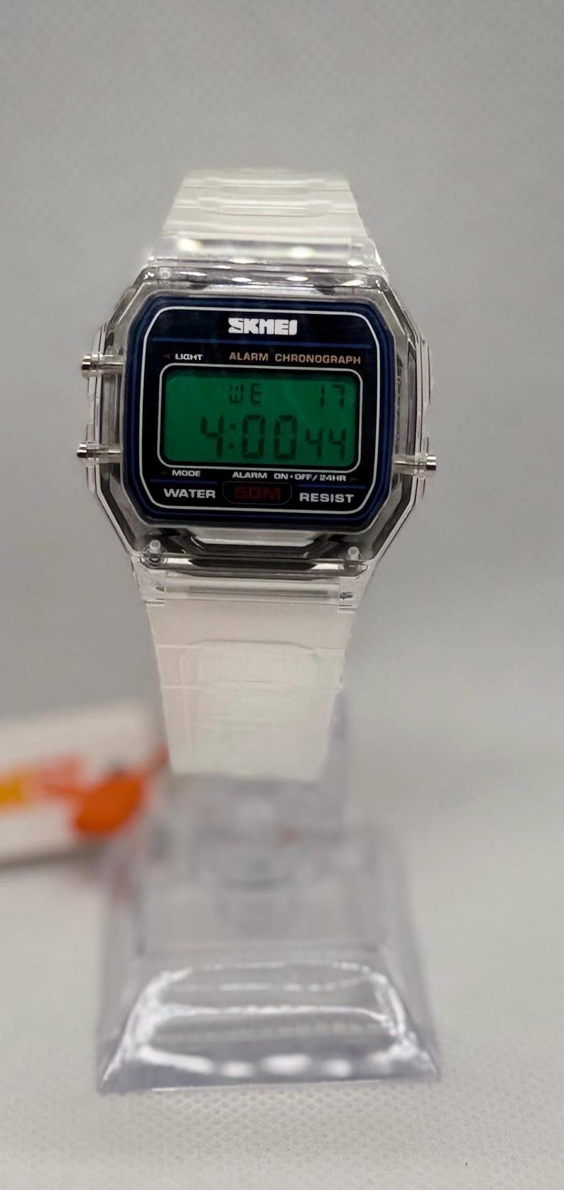 K&ouml;nnte beinhalten: Transparente Digitaluhr mit rechteckigem Zifferblatt. Die Uhr zeigt die Zeit 4:00:44 auf einem gr&uuml;nen Bildschirm an. Sie hat ein transparentes Kunststoffarmband und ein schwarz-blaues Zifferblatt mit der Marke "SKMEI".
