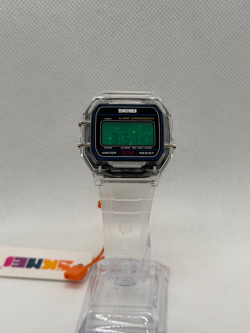 K&ouml;nnte beinhalten: Transparente Digitaluhr mit rechteckigem Zifferblatt und transparentem Armband. Das Zifferblatt hat einen schwarzen Rand mit dem Text "SKMEI" und eine gr&uuml;ne Digitalanzeige. Armband und Uhrengeh&auml;use sind transparent.