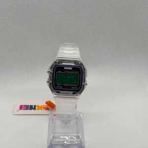 K&ouml;nnte beinhalten: Transparente Digitaluhr mit rechteckigem Zifferblatt und transparentem Armband. Das Zifferblatt zeigt die Uhrzeit in gr&uuml;nen Ziffern an. Der Markenname "SKMEI" ist auf dem Zifferblatt sichtbar. Die Uhr wird auf einem transparenten Kunststoffst&auml;nder pr&auml;sentiert.