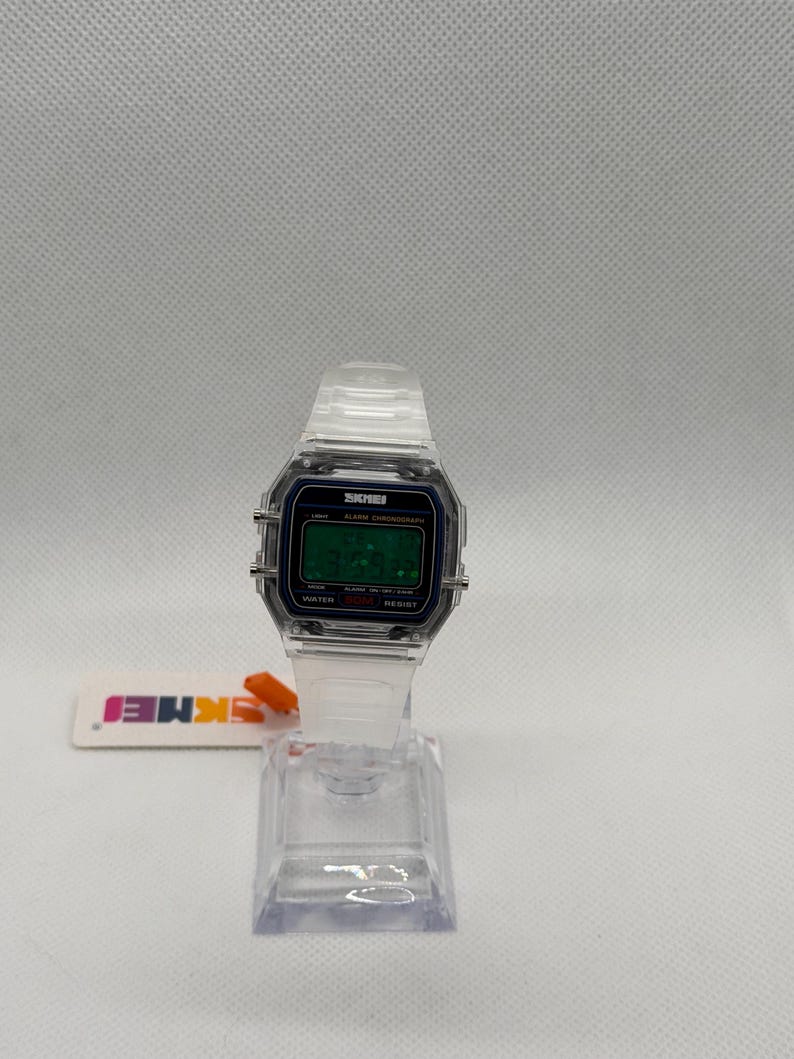 K&ouml;nnte beinhalten: Transparente Digitaluhr mit rechteckigem Zifferblatt und transparentem Armband. Das Zifferblatt zeigt die Uhrzeit in gr&uuml;nen Ziffern an. Der Markenname "SKMEI" ist auf dem Zifferblatt sichtbar. Die Uhr wird auf einem transparenten Kunststoffst&auml;nder pr&auml;sentiert.