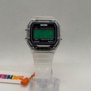 K&ouml;nnte beinhalten: Transparente Digitaluhr mit quadratischem Zifferblatt und wei&szlig;em Armband. Das Zifferblatt zeigt die Uhrzeit in gr&uuml;nen Ziffern an. Die Uhr tr&auml;gt den Markennamen "SKMEI" auf dem Zifferblatt.