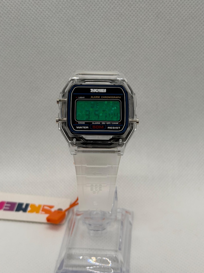 K&ouml;nnte beinhalten: Transparente Digitaluhr mit quadratischem Zifferblatt und wei&szlig;em Armband. Das Zifferblatt zeigt die Uhrzeit in gr&uuml;nen Ziffern an. Die Uhr tr&auml;gt den Markennamen "SKMEI" auf dem Zifferblatt.