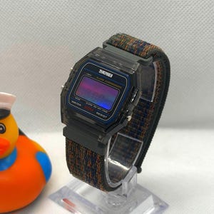 May include: A digital wristwatch with a translucent dark gray plastic case and a multicolored woven fabric strap. The watch face features the text SKMEI, ALARM CHRONOGRAPH, and WATER 50M RESIST, with a purple and blue gradient digital display screen.