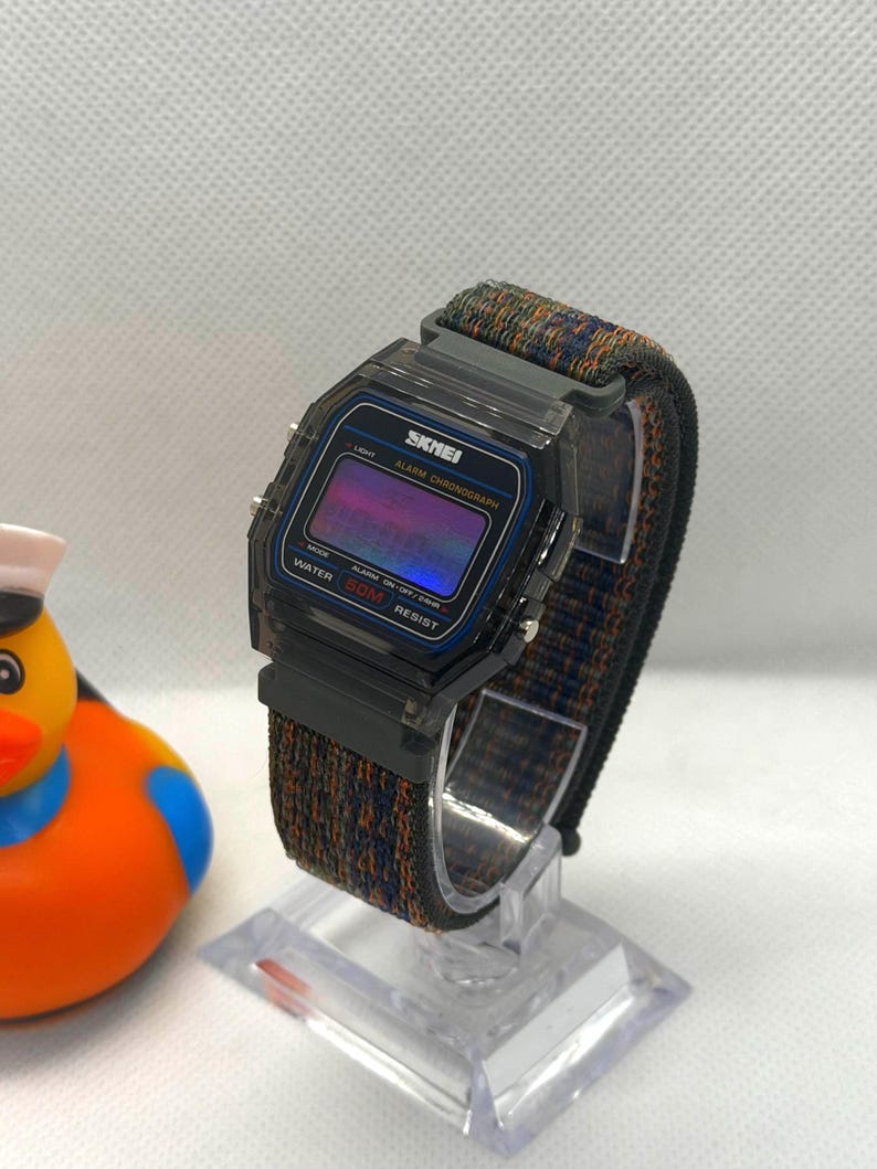 May include: A digital wristwatch with a translucent dark gray plastic case and a multicolored woven fabric strap. The watch face features the text SKMEI, ALARM CHRONOGRAPH, and WATER 50M RESIST, with a purple and blue gradient digital display screen.