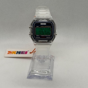 K&ouml;nnte beinhalten: Transparente Digitaluhr mit rechteckigem Zifferblatt und passendem transparentem Armband. Das Zifferblatt zeigt die Uhrzeit und tr&auml;gt den Markennamen "SKMEI". Die Uhr wird auf einem transparenten Kunststoffst&auml;nder pr&auml;sentiert.