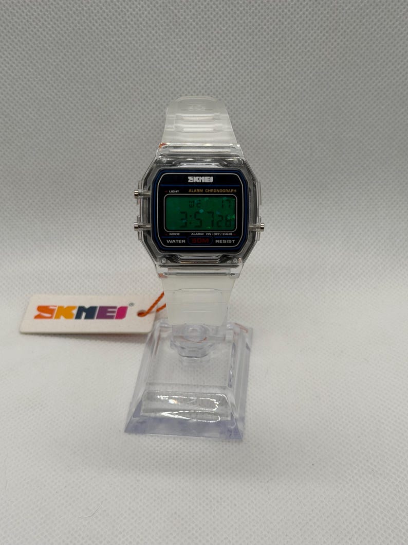 K&ouml;nnte beinhalten: Transparente Digitaluhr mit rechteckigem Zifferblatt und passendem transparentem Armband. Das Zifferblatt zeigt die Uhrzeit und tr&auml;gt den Markennamen "SKMEI". Die Uhr wird auf einem transparenten Kunststoffst&auml;nder pr&auml;sentiert.
