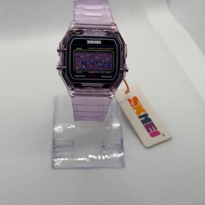 Puede incluir: Un reloj digital lila transparente con una correa a juego. La esfera del reloj muestra la hora y otras funciones. La marca "SKMEI" es visible en la esfera y en la etiqueta adjunta.