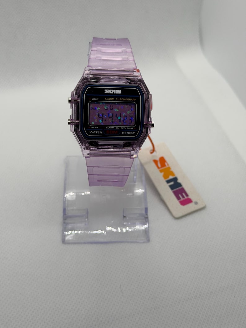 Puede incluir: Un reloj digital lila transparente con una correa a juego. La esfera del reloj muestra la hora y otras funciones. La marca "SKMEI" es visible en la esfera y en la etiqueta adjunta.