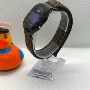 May include: A digital wristwatch with a black rectangular case and a dark blue screen, featuring a textured nylon strap in shades of olive green, navy blue, and orange. The watch is mounted on a clear plastic display stand next to a rubber duck.