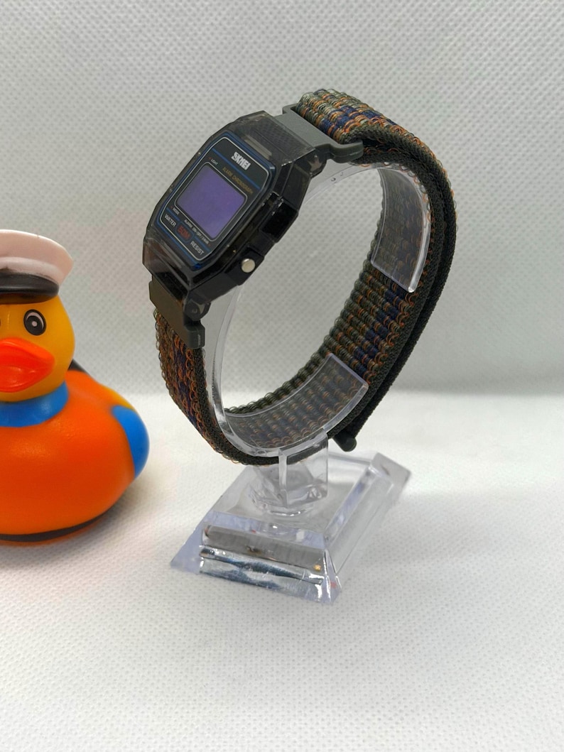 May include: A digital wristwatch with a black rectangular case and a dark blue screen, featuring a textured nylon strap in shades of olive green, navy blue, and orange. The watch is mounted on a clear plastic display stand next to a rubber duck.