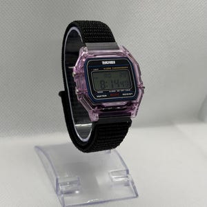 May include: A digital watch with a black nylon band and a transparent pink case. The watch face displays the time, date, and other functions. The watch is labeled "SKMEI" and is water-resistant.