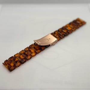 Op de afbeelding: Een horlogeband met een schildpadpatroon in bruine en amberkleurige tinten. De band heeft een roségouden sluiting. De rechthoekige schakels zijn verbonden om een flexibele band te vormen.