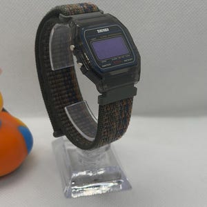 May include: A digital wristwatch with a translucent black plastic case and a rectangular face featuring the brand name SKMEI. The watch has a dark, multi-colored woven fabric strap with blue and orange threads and silver-tone side buttons.