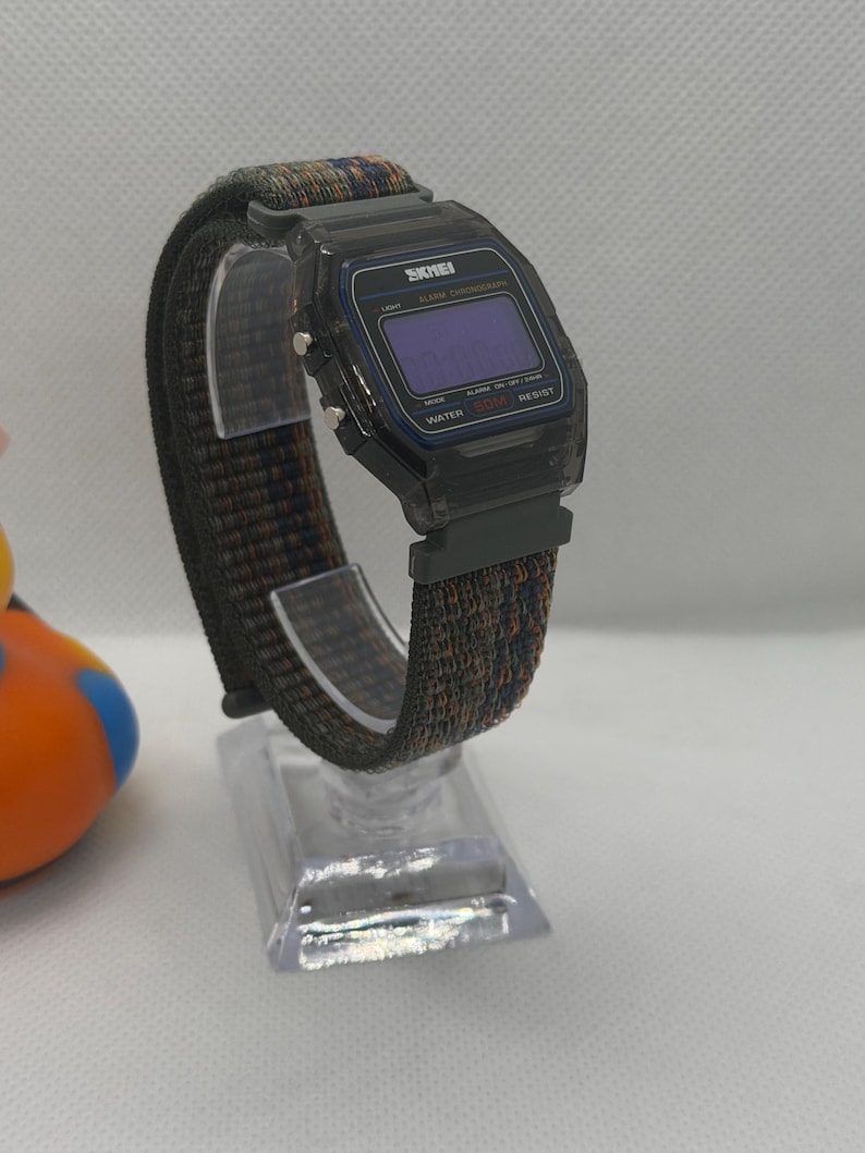 May include: A digital wristwatch with a translucent black plastic case and a rectangular face featuring the brand name SKMEI. The watch has a dark, multi-colored woven fabric strap with blue and orange threads and silver-tone side buttons.